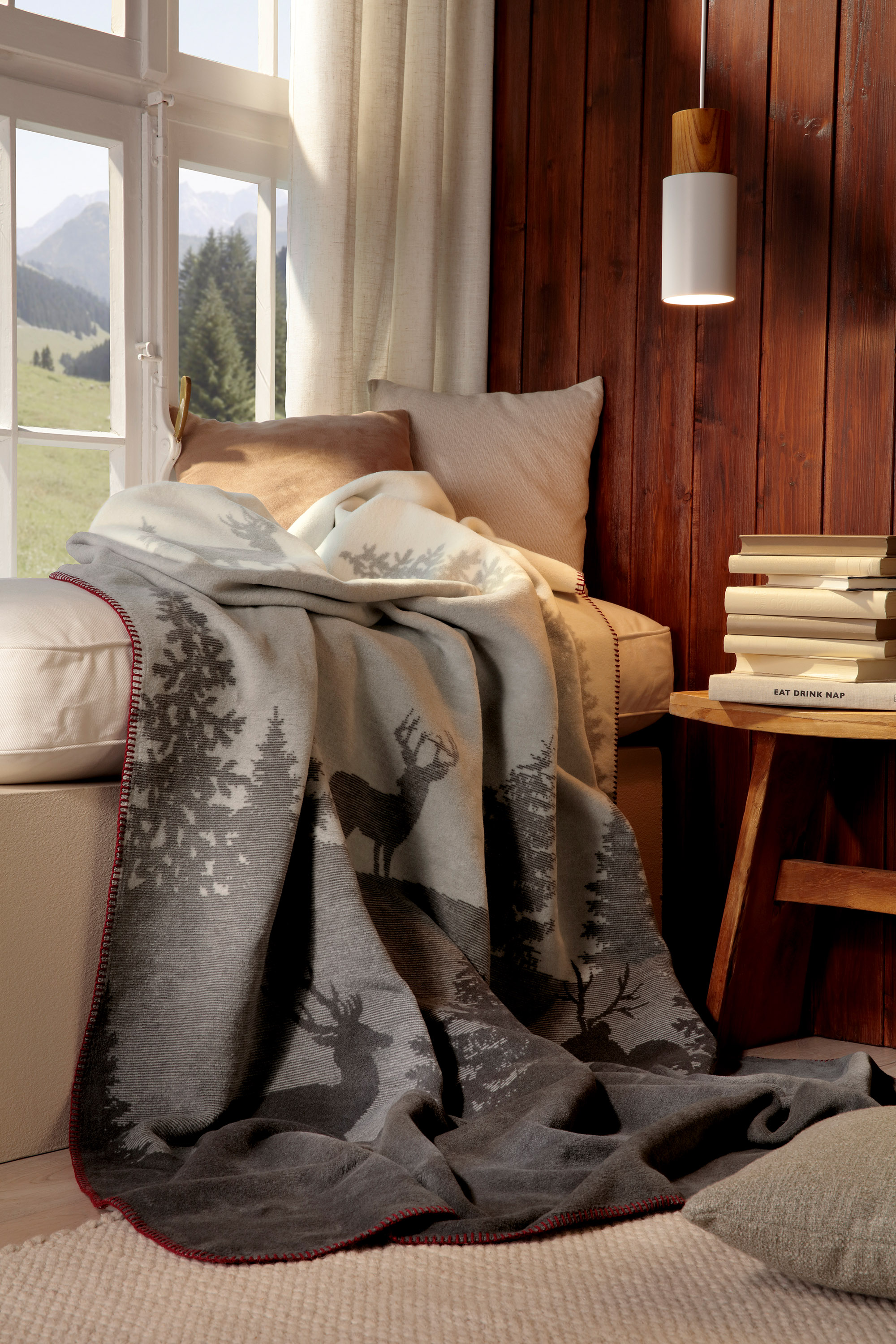 Wohndecke Deerlight auf Sofa liegend vor einem Fenster mit Bücher im Vordergrund
